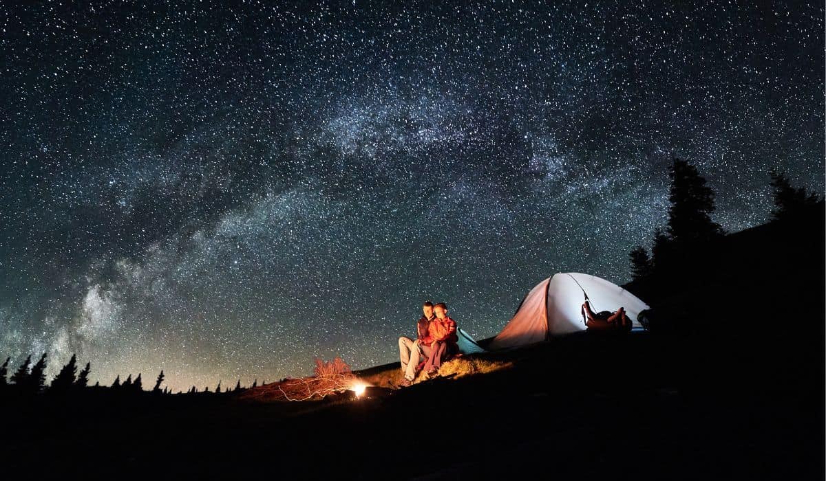 Night camping. Romantic couple tourists have a rest at a campfire near illuminated tent under amazing night sky full of stars