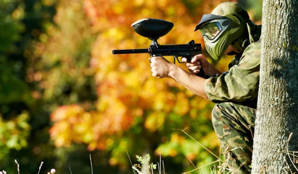 a photograph of an individual playing paintball hiding near a tree