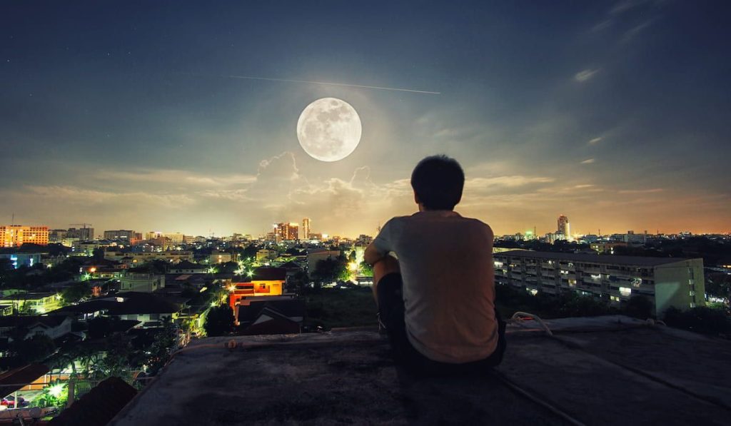 man sitting alone at night on his rooftop, seeing the whole city and watching the full moon