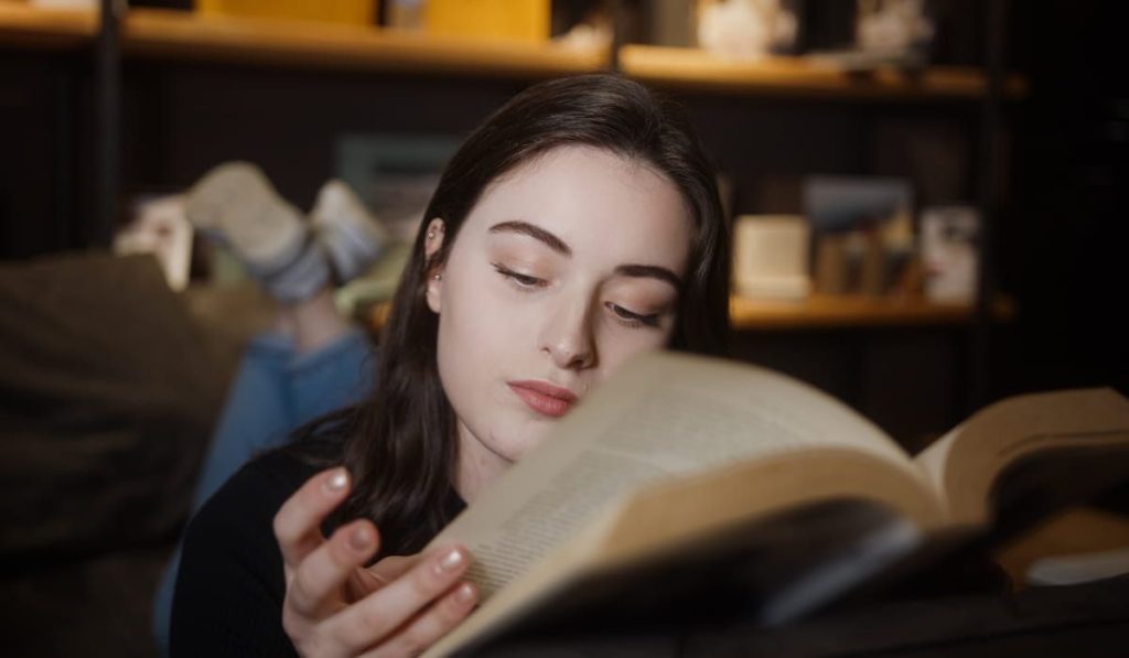 young woman lying on sofa at home reading novel 