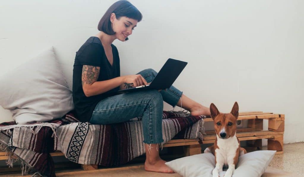 Pretty woman writes a blog on laptop, her best friend dog puppy lays next to her on pillow
