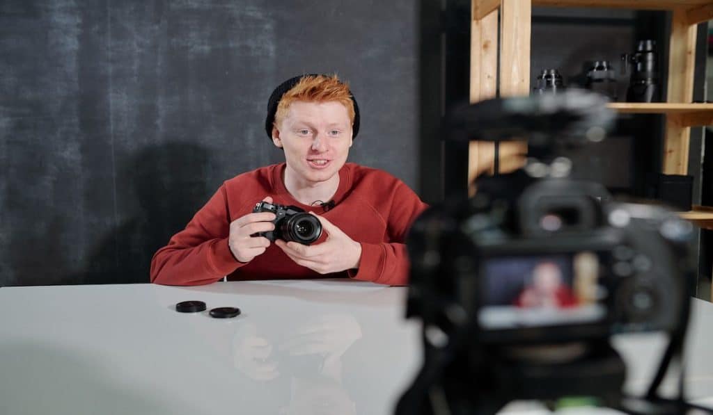 Cheerful young vlogger by the desk holding new photocamera while presenting it in front of his camera