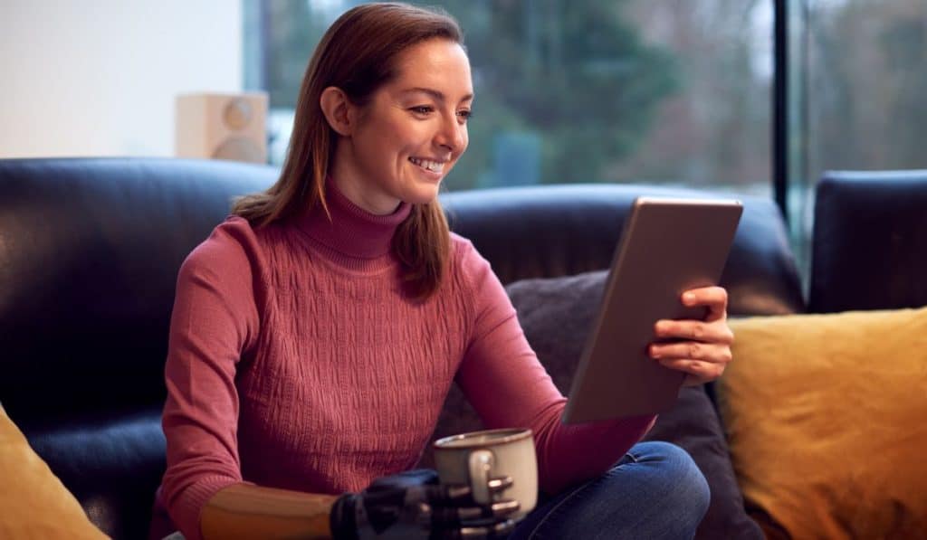 Woman with prosthetic arm hand at home reading from her tablet while holding cup of coffee