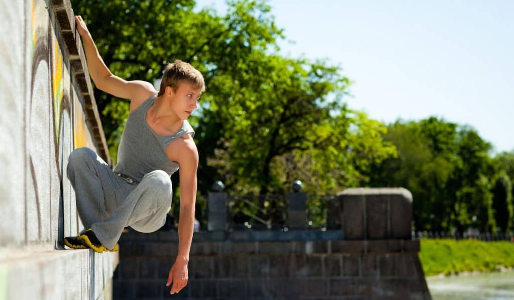 Young man in sportswear practicing parkour outside on stone wall on clear summer day