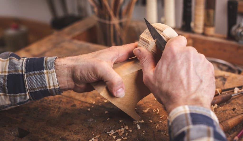 Hands of a man using a knife to carve a small piece of wood on a workbench
