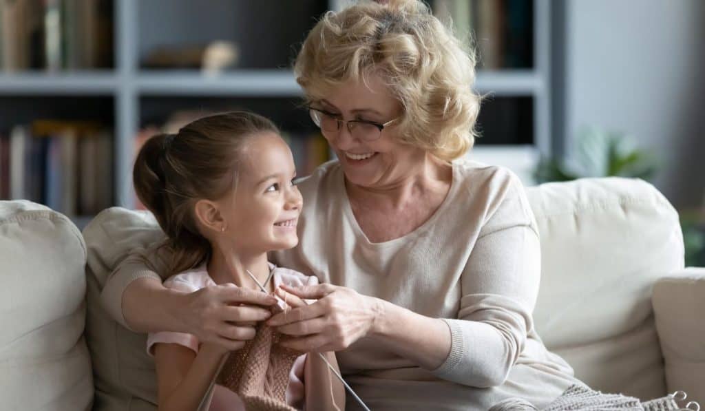 Smiling loving mature grandmother wearing glasses teaching little granddaughter knitting