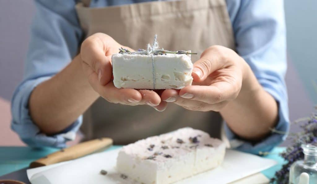 Woman holding hand made soap bar with lavender flowers at light blue wooden table, closeup
