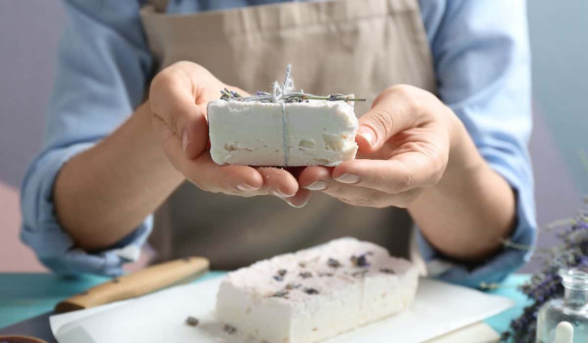 Woman holding hand made soap bar with lavender flowers at light blue wooden table closeup