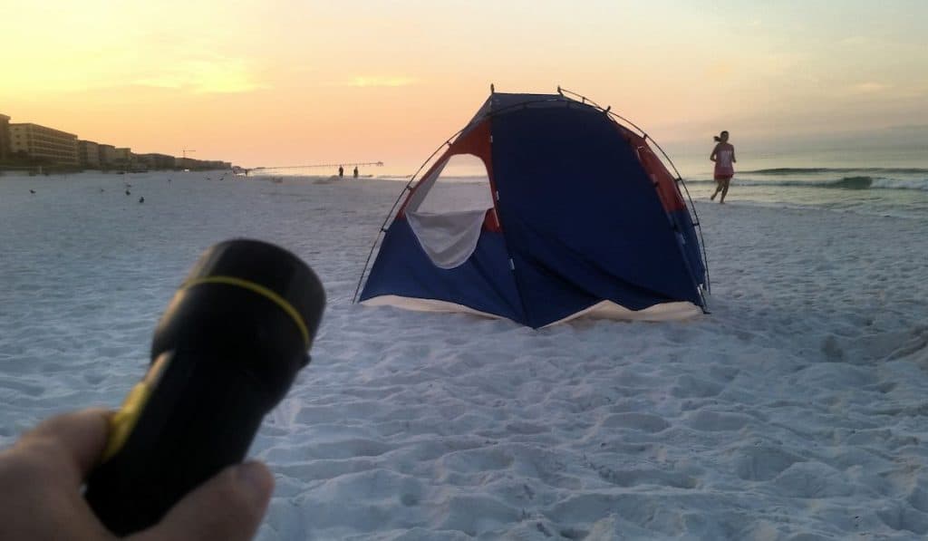 A hand holding a flashlight pointing a tent on the beach