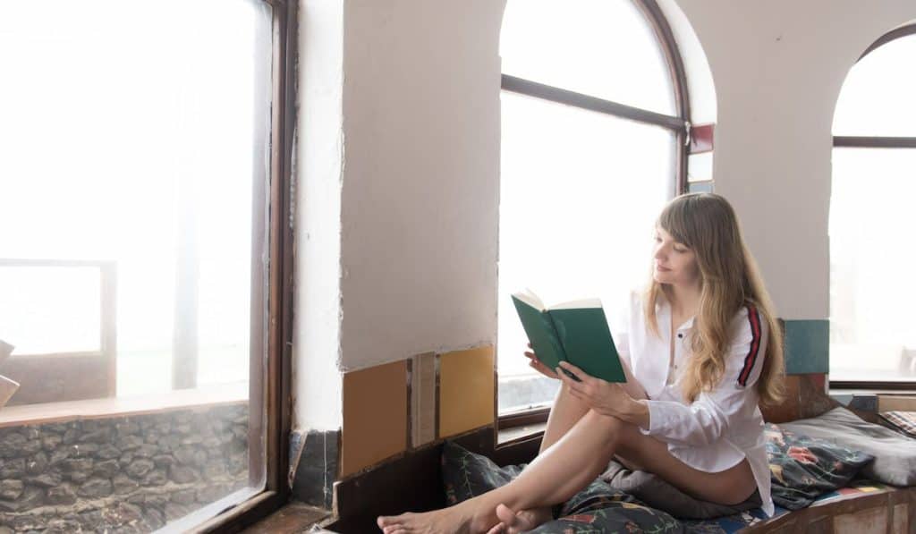 barefoot female in shirt sitting on pillows near window and reading interesting book on sunny day