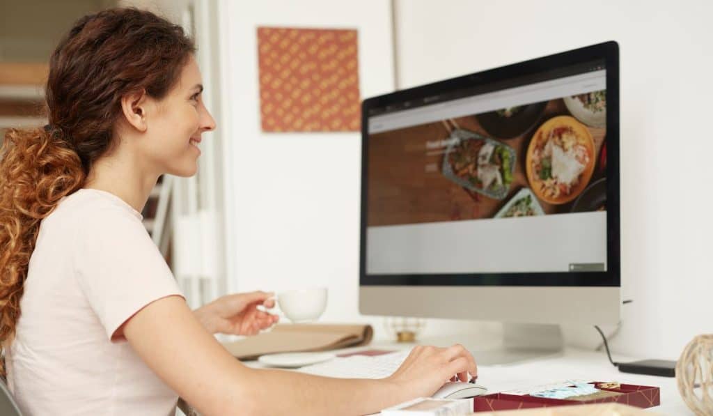 female web designer with curly hair sitting at table and drinking coffee while working on website in office