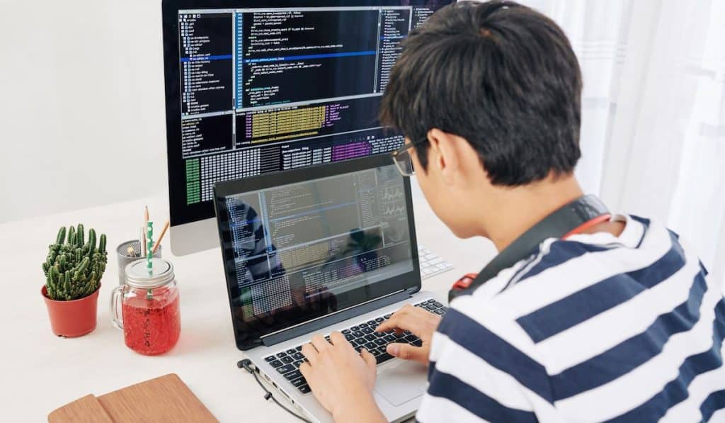 teenager working on laptop, programming code on computer screen in background