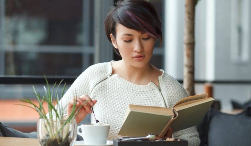 woman sitting at table stirring up coffee with spoon and reading book in café