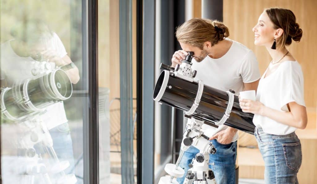 Young couple watching with telescope standing near the window in the modern house 