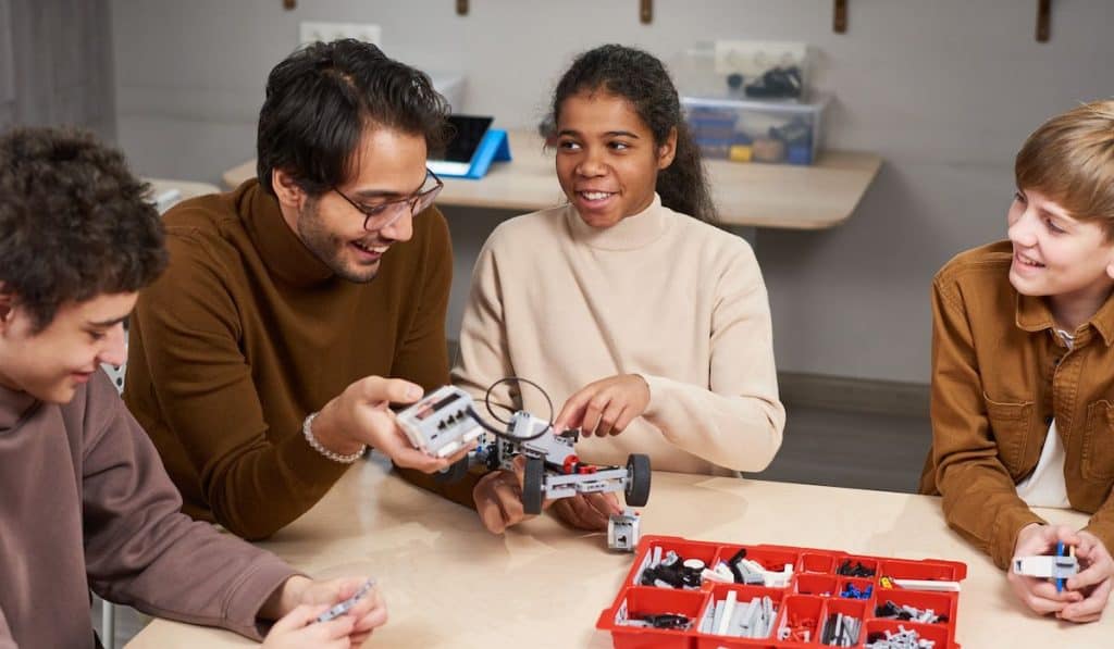 school children with young male teacher investigating electronic constructor at the table at robotics lesson