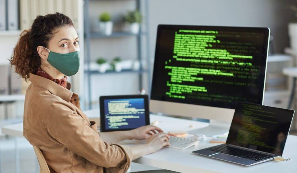  young programmer in protective mask looking at camera while sitting at her workplace 